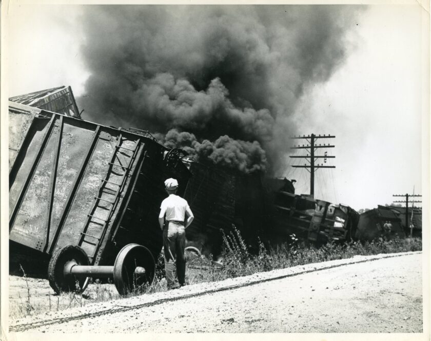 Un unidentified male stands near a set of wheels detached from a freight car. The railroad cars are derailed and on fire. Large clouds of black smoke fill the air.