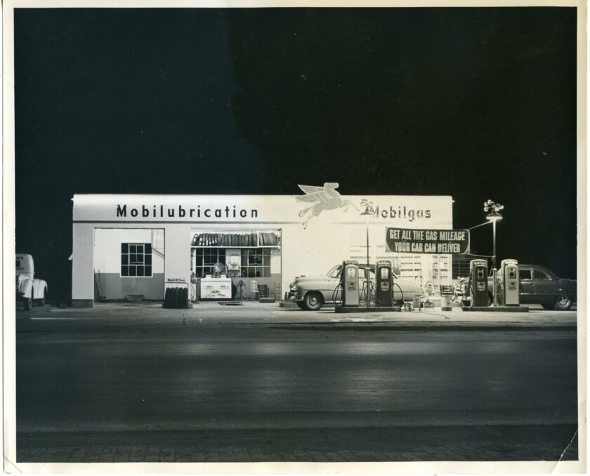 In this black-and-white image shot at night, the Mobilubrication station, a brightly lit white building, stands along an empty street.