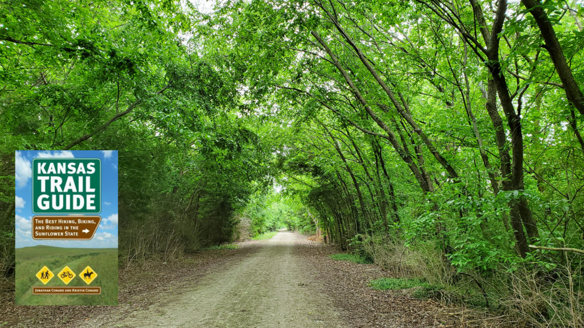 Green-leafed trees arch over a gravel trail. The cover of the book Kansas Trail Guide is highlighted in the lower left corner.