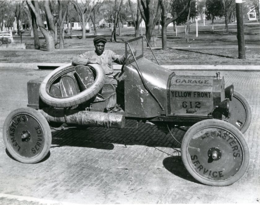 An African American man sits behind the wheel of a 1920s race car, which is parked in front of City Park. Trees and a pedestrian bridge are visible in the background. The man rests his arm on a tire. Printed on the car are "Lemaster's Garage Yellow Front" and "Lemaster's service."