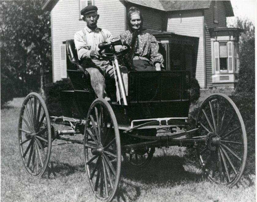 And elderly man and woman sit in a very early automobile, which is parked in front of a house.