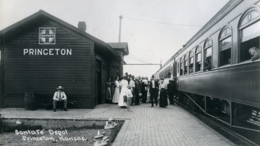 A train is pulled in to a Santa Fe depot. the Santa Fe logo and "Princeton" are visible under the gable of the depot's roof. People are gathered on the platform between the depot and the train. Passengers can be seen inside the train.