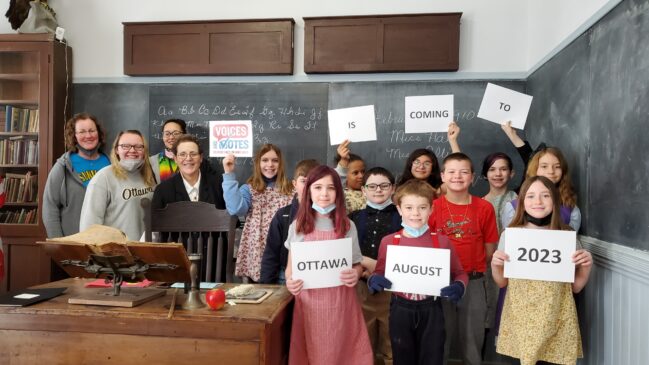 School children hold signs that say "Voices and Votes" is coming to Ottawa in 2023