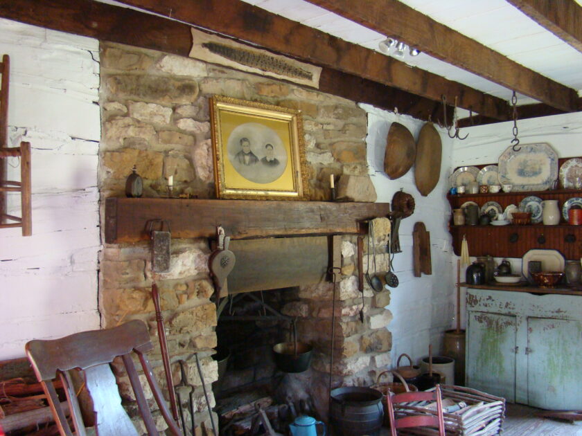 Image shows a stone fireplace. A large gold-framed wedding photo of a couple rests on a mantel. The fireplace is surrounded by household items common in the 1800s.