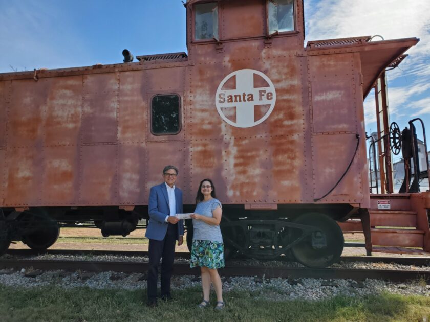 A man hands a woman a check while standing in front of a caboose.