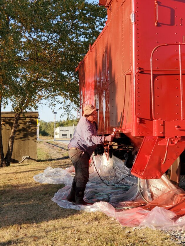 A man paints the side of a caboose with a paint sprayer