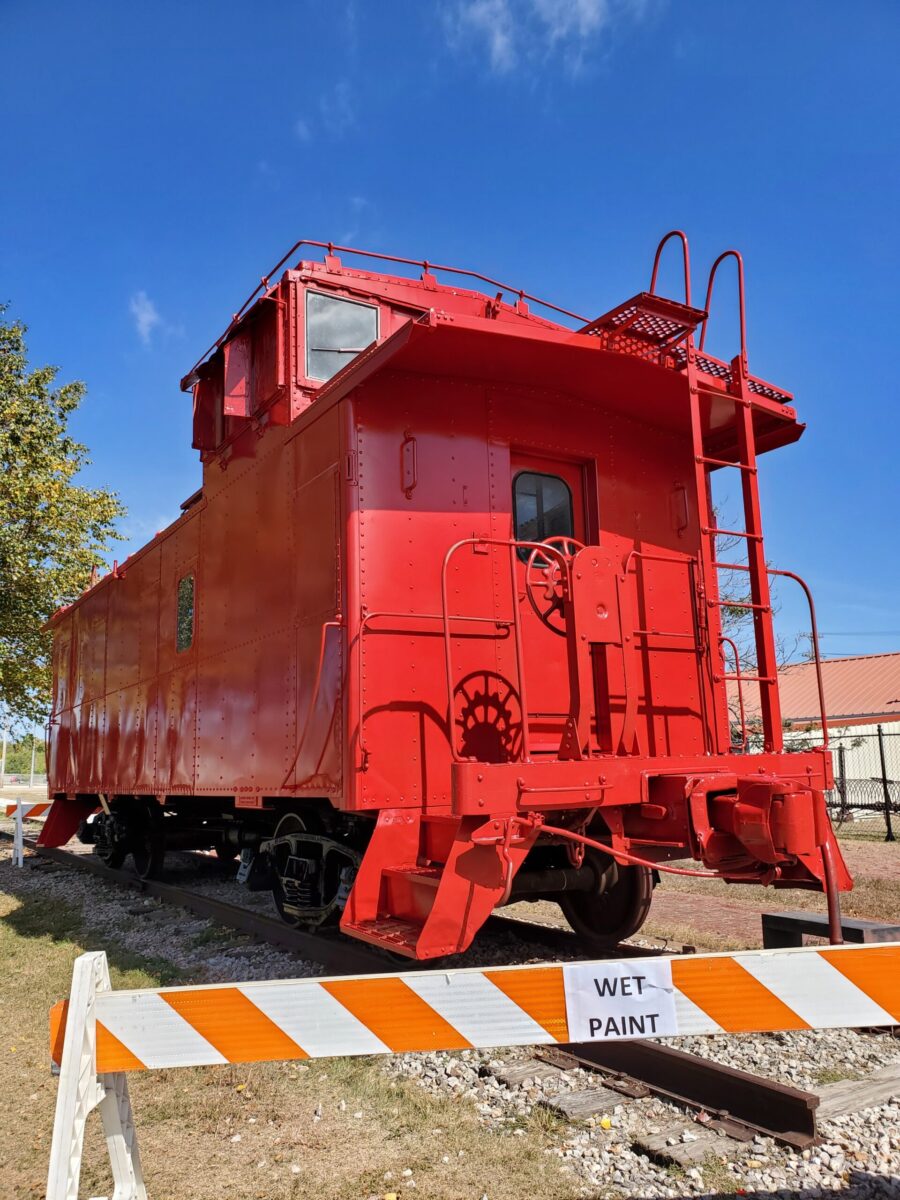 Brightly painted caboose. A "Wet Paint" sign hangs from a barrier in front of it.
