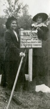 Two women wearing 1910s-era suits and hats stand by a sign that says "WARNING; Trespassing upon the tracks and Right of Way is positively forbidden.:" One woman holds a pitchfork.